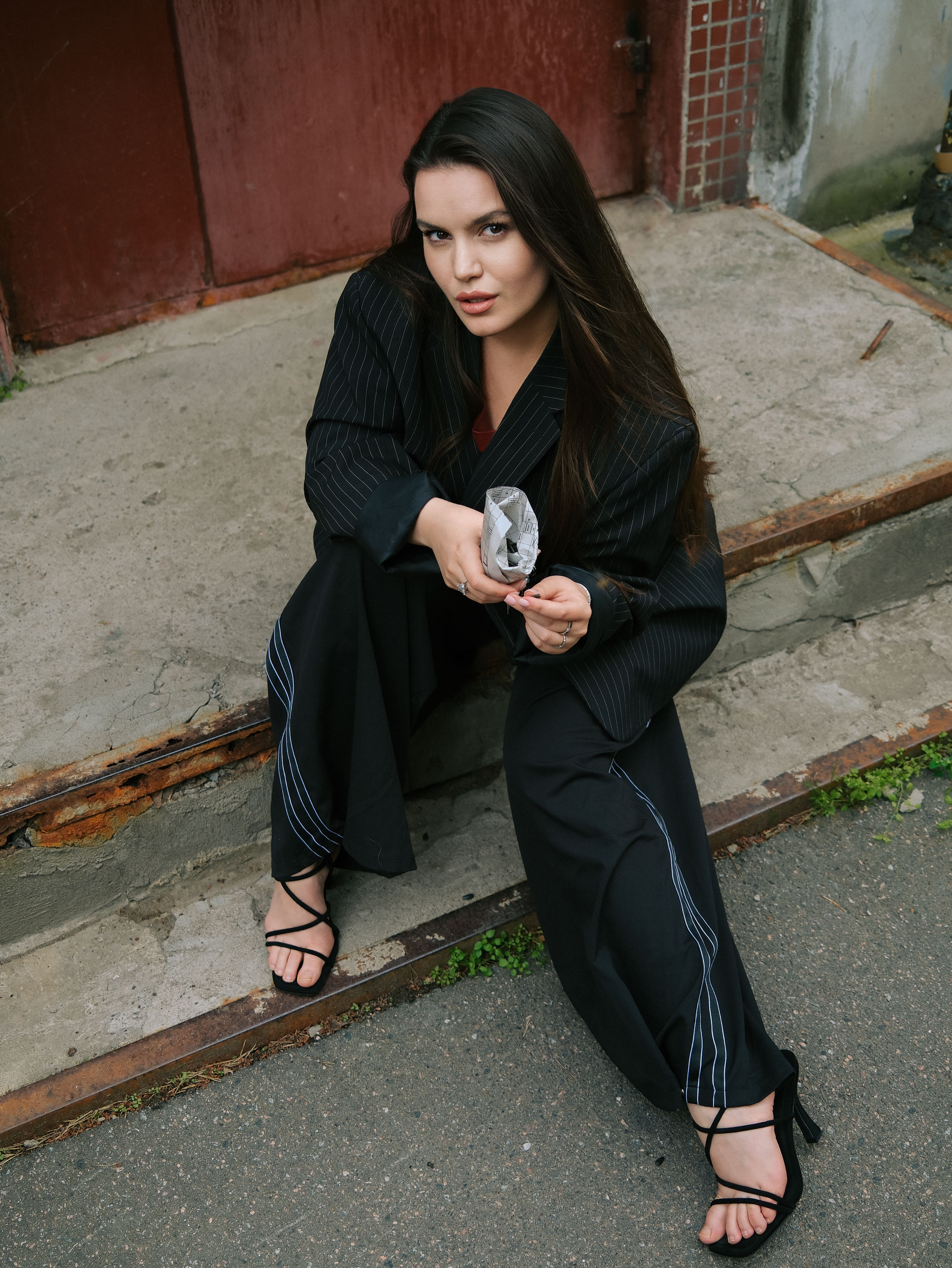 Fashionable woman in a black pantsuit seated on rustic outdoor steps on a city street.