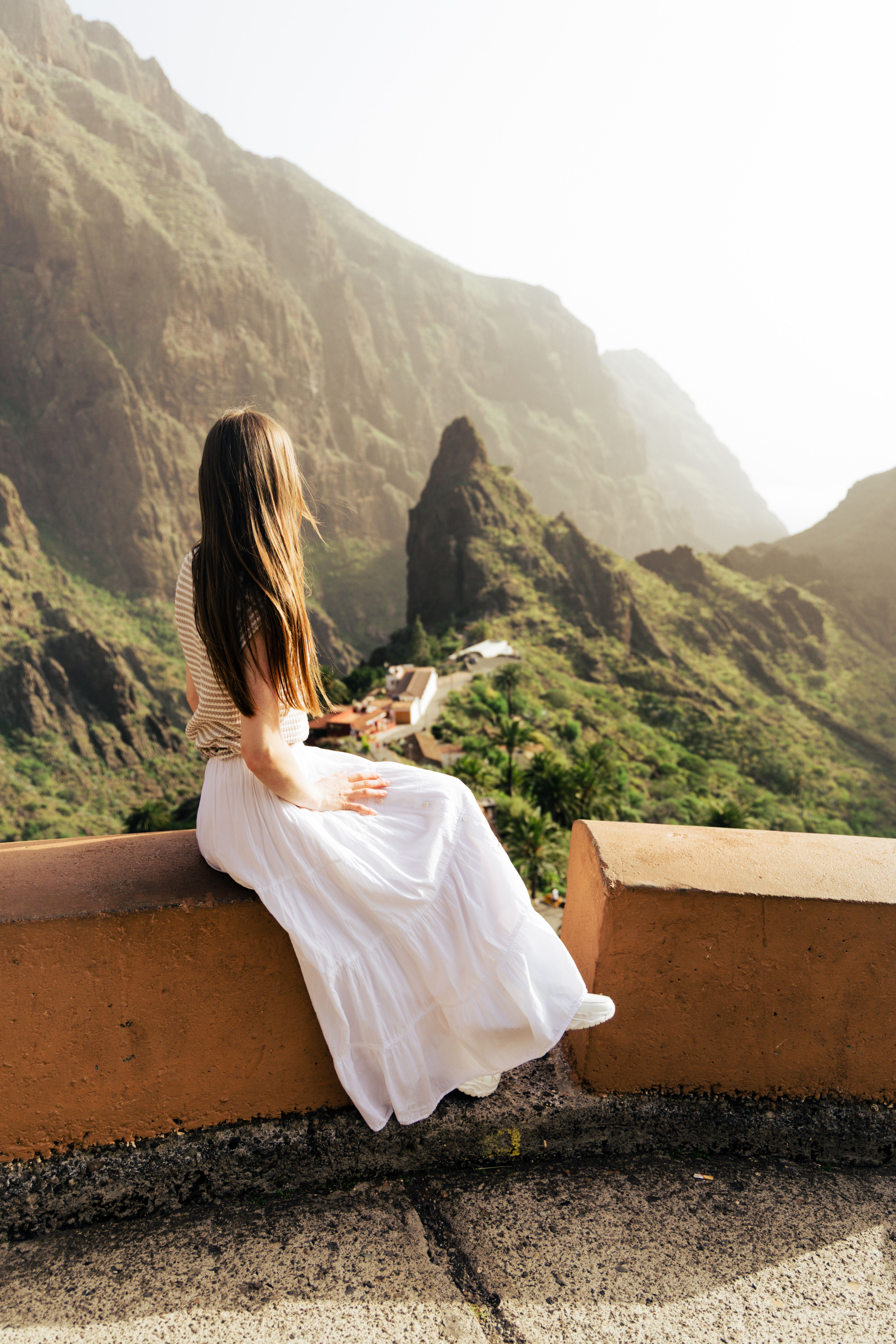 Young woman in a white dress sits overlooking Masca Valley, a stunning natural landscape in Spain.
