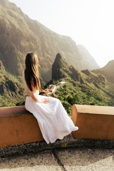 Young woman in a white dress sits overlooking Masca Valley, a stunning natural landscape in Spain.