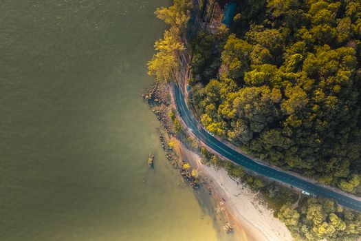 High-angle aerial shot of a winding coastal road flanked by lush forest and tranquil shoreline.