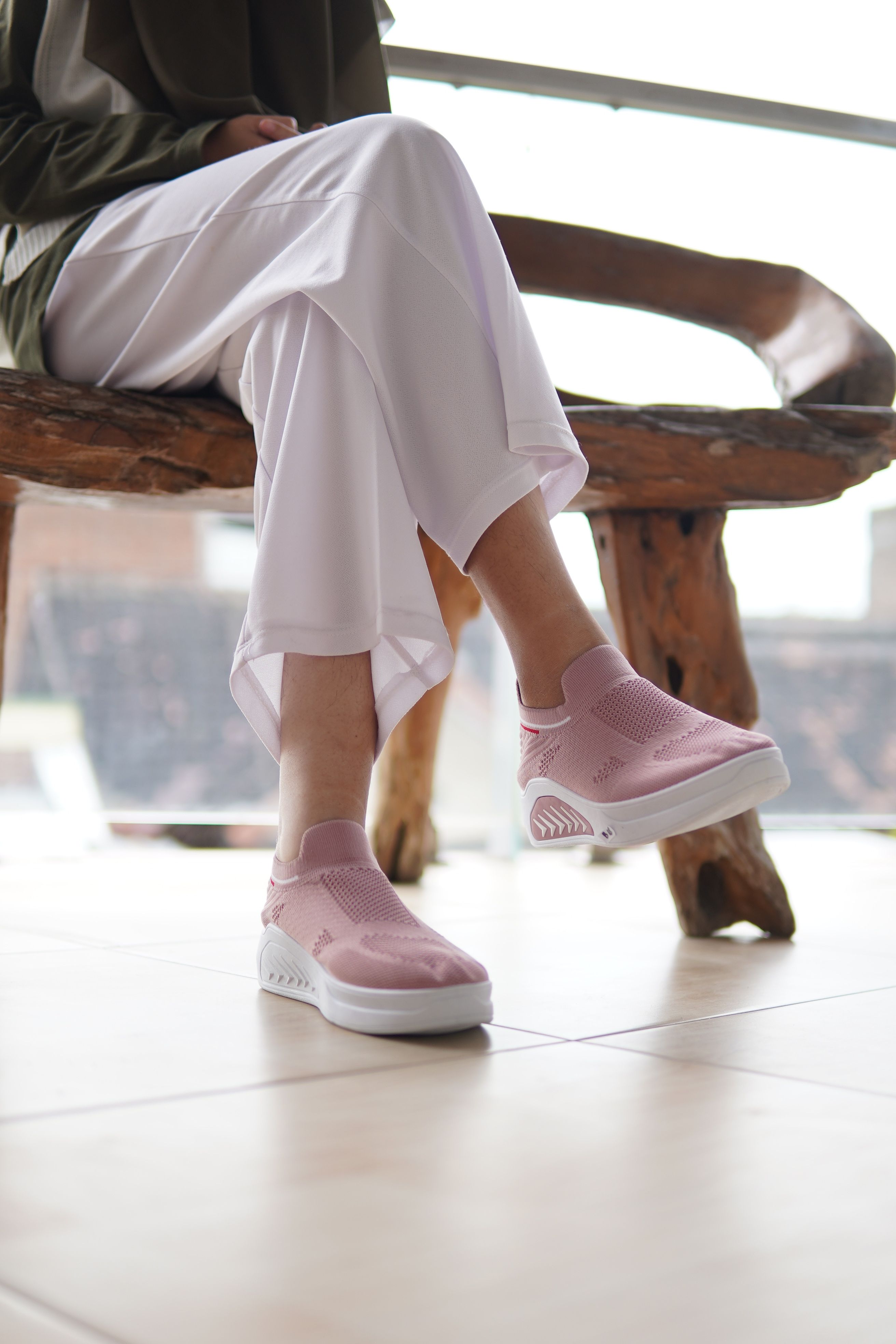 A person sitting on a rustic chair indoors wearing pink sneakers and white pants.