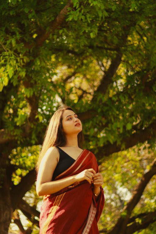 Free Young woman in red sari under lush tree in sunlight, serene expression. Stock Photo
