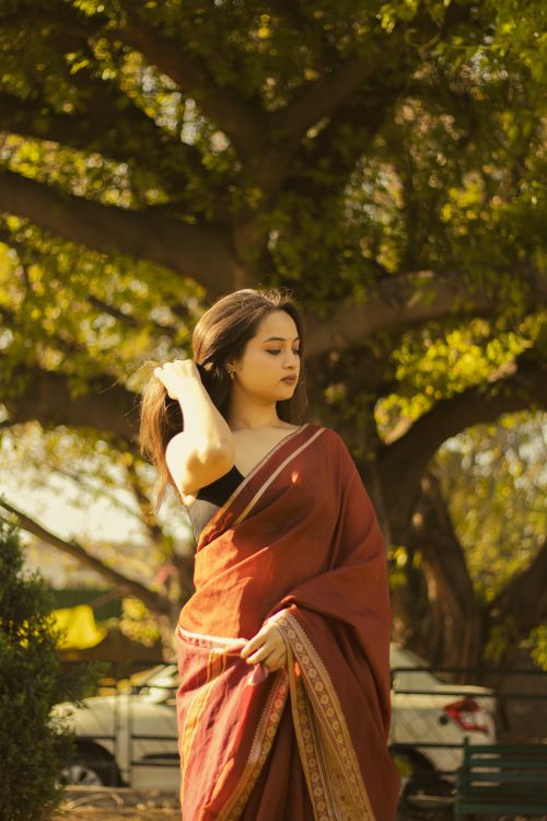 Free A woman in a red sari stands outdoors, illuminated by warm sunlight, under a large tree. Stock Photo