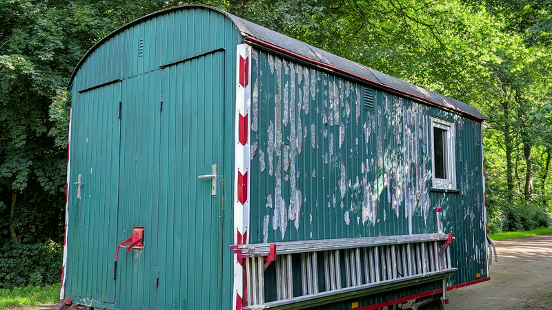 An old green wooden caravan on wheels with peeling paint among trees
