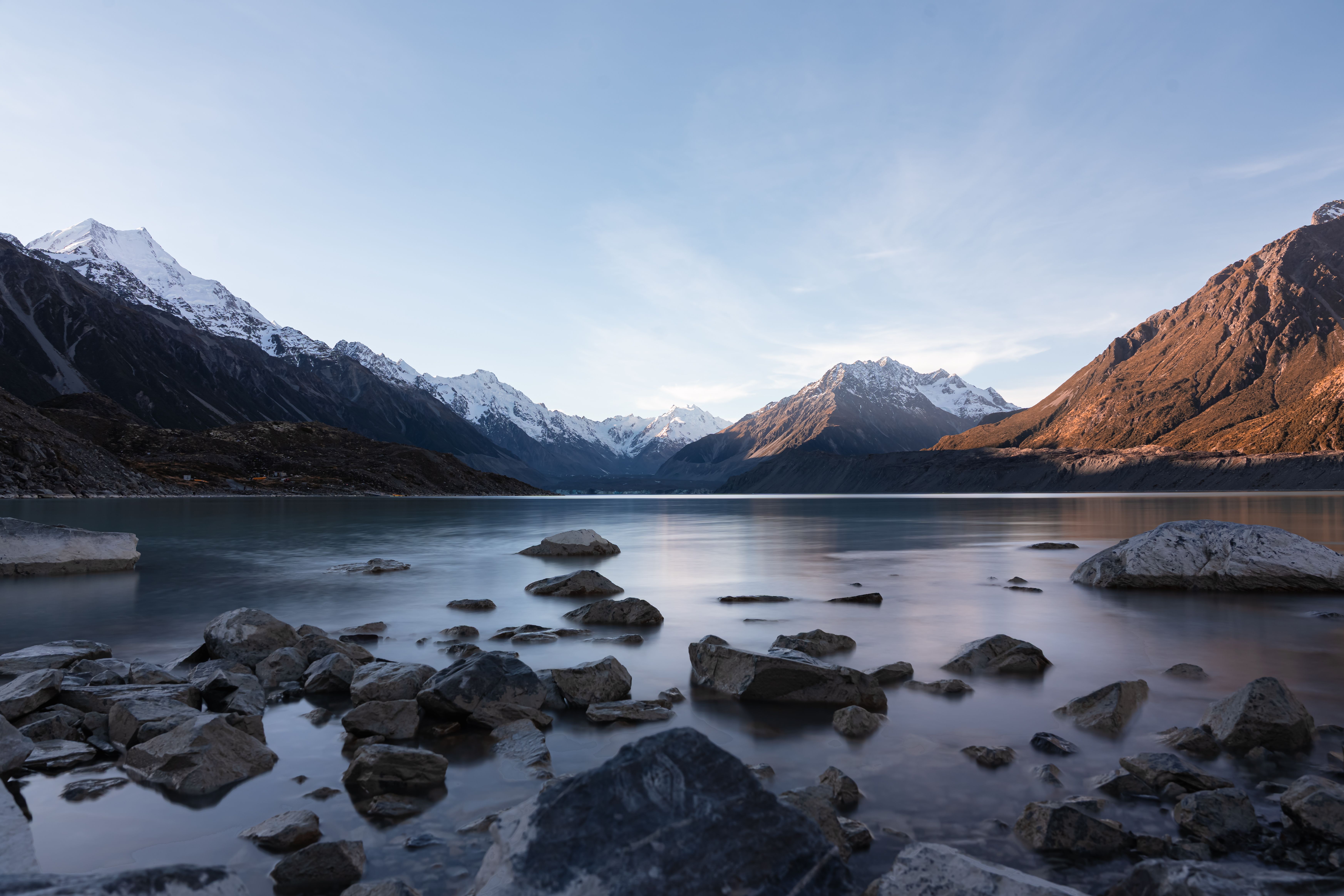 Tranquil mountain lake scene with rocky shore and snow-capped peaks at sunset, reflecting nature's beauty.