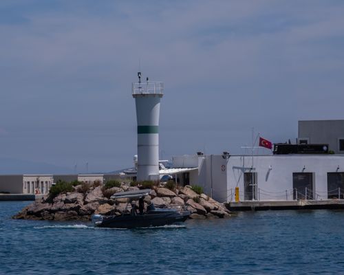 Free Picturesque marina scene in Foça, İzmir with a lighthouse, yacht, and Turkish flag under a clear blue sky. Stock Photo