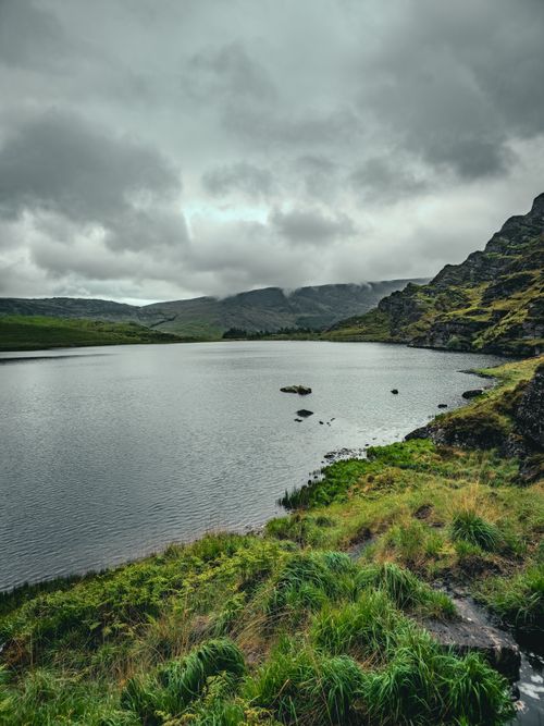 Free A serene mountain lake surrounded by lush greenery under a cloudy sky, captured during the day. Stock Photo
