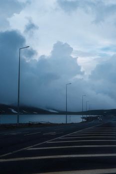 Moody clouds hover over a quiet road by a lake, creating a dramatic atmosphere.