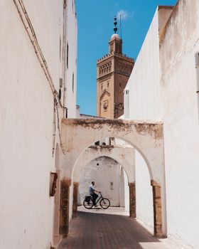 Cyclist rides through an archway in a narrow street of a historic medina with a tall mosque tower visible in the background.