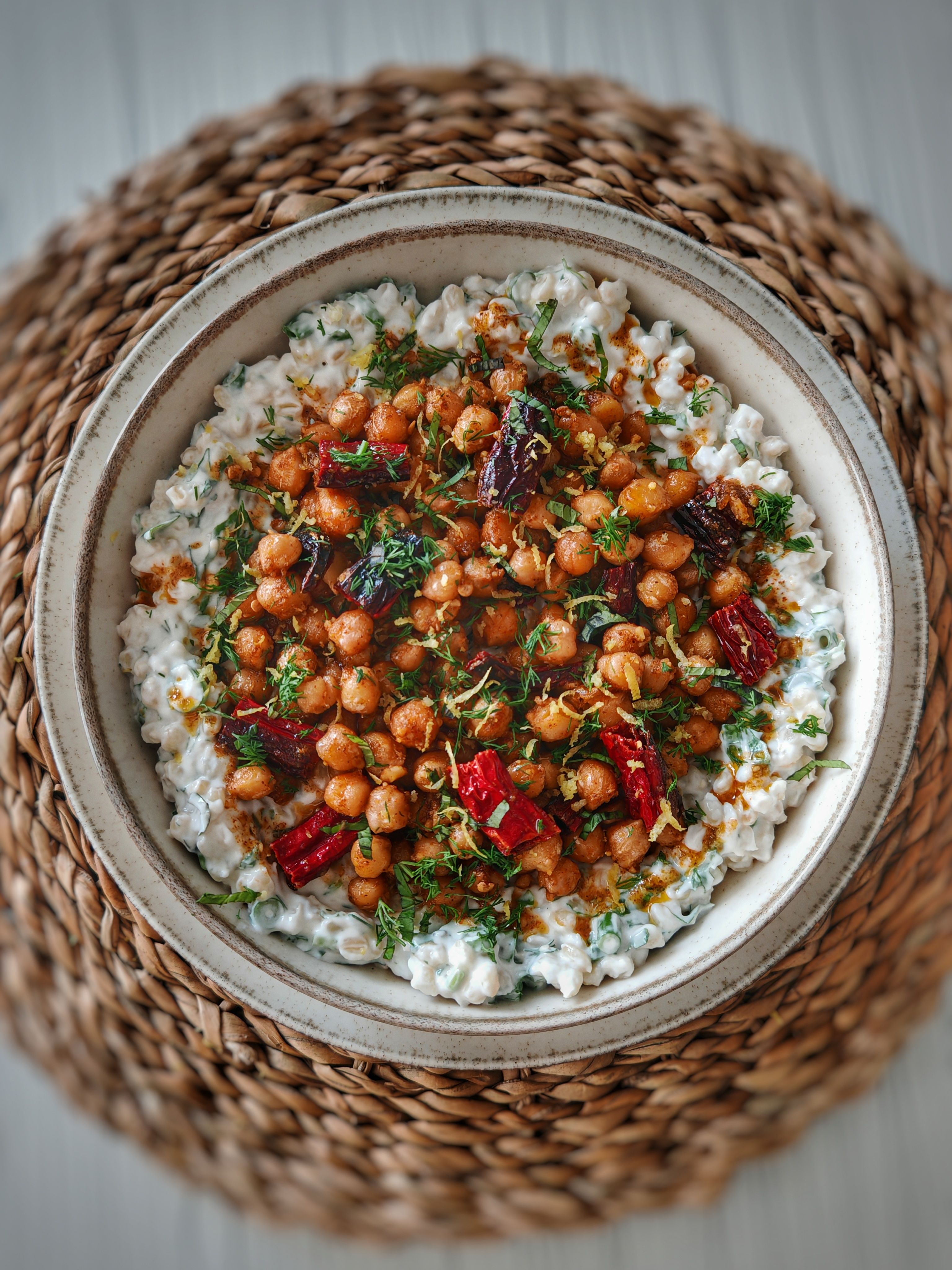 A bowl of glossy harissa-coated noodles topped with crispy roasted chickpeas, charred tomatoes, fresh cilantro, toasted almonds, and a dollop of white yogurt, with a lemon wedge on the side.