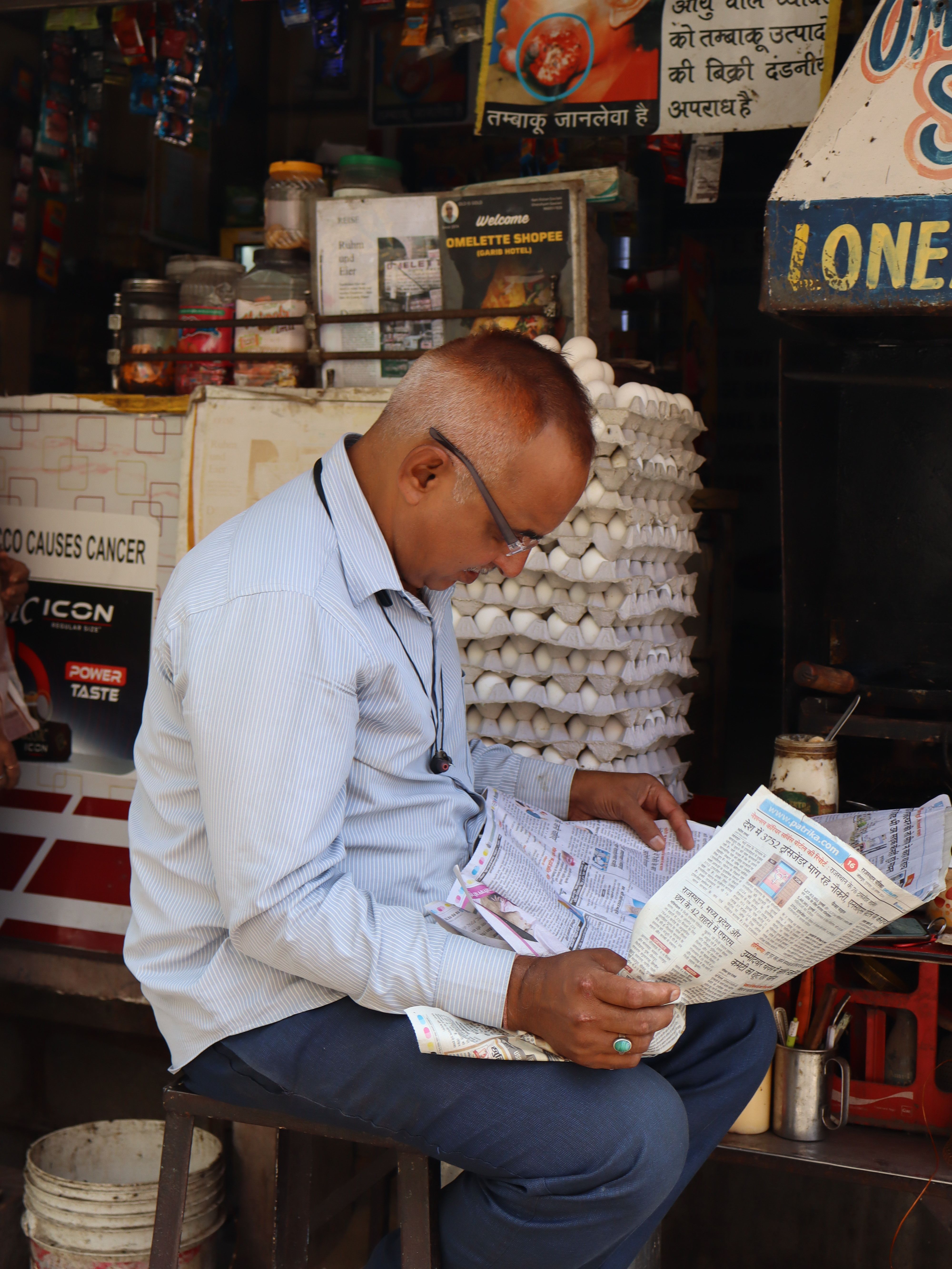Candid street portrait of a man engrossed in reading a newspaper at a bustling market stall in Jodhpur, India.
