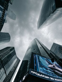 Dramatic angle view of modern skyscrapers under a cloudy sky with a prominent digital billboard.