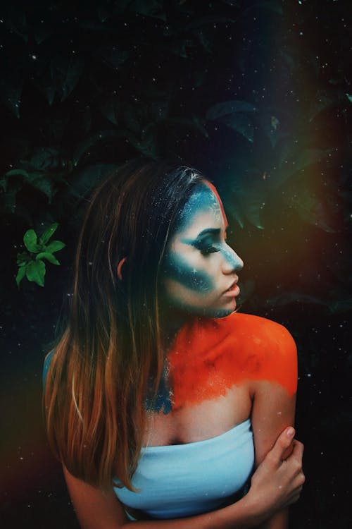 From above of young slim woman with blue paint on face and orange paint on shoulders representing cold and hot weather standing with closed eyes near tree branch with leaves