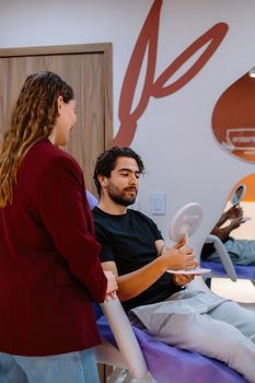 A male client receives a consultation at a clinic, reviewing reflections in a hand mirror.