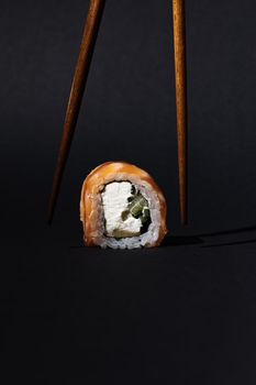 Close-up of salmon sushi roll with chopsticks on black surface.