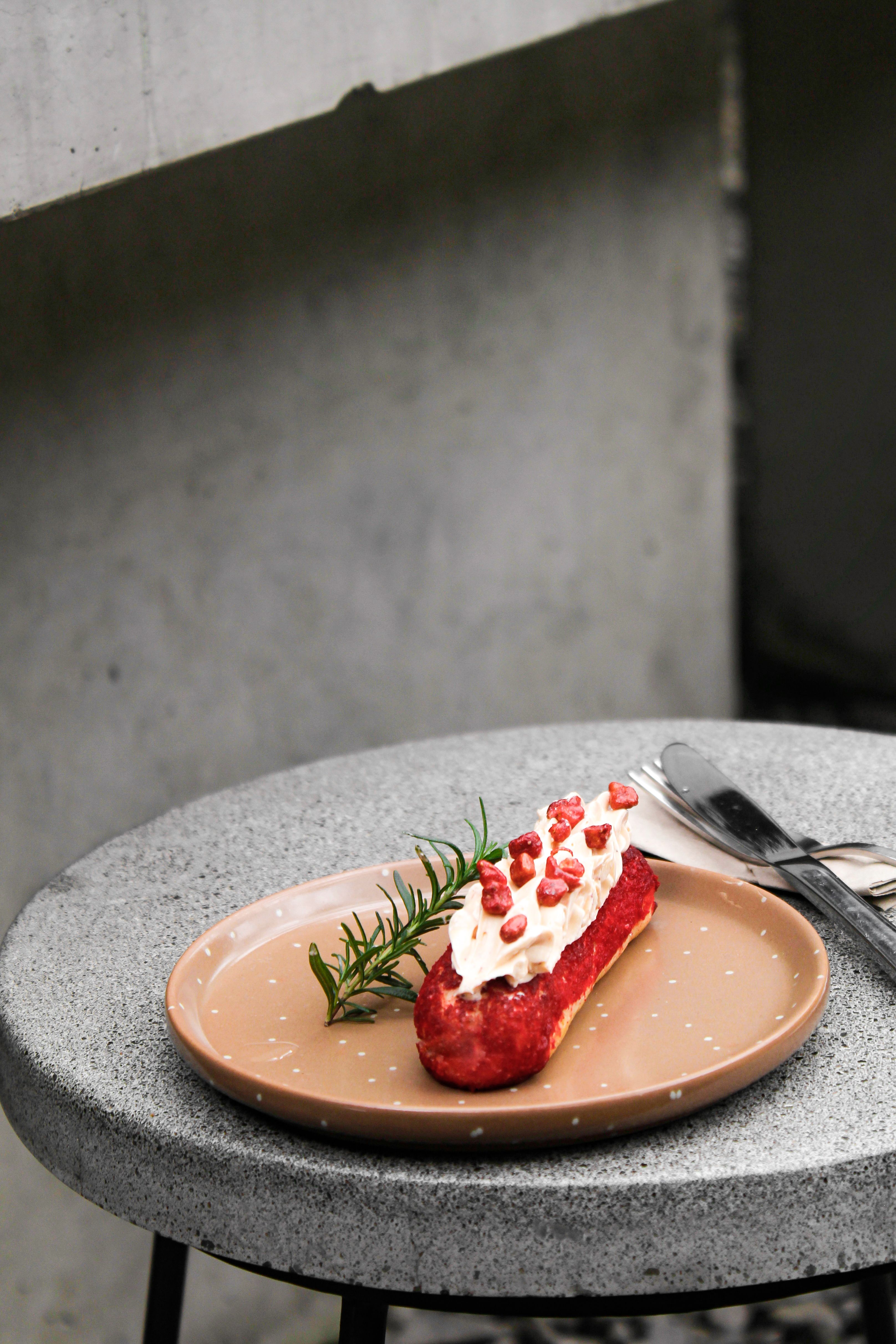 Tantalizing red eclair topped with cream and pomegranate seeds on a plate.