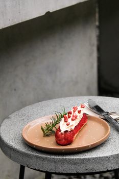 Tantalizing red eclair topped with cream and pomegranate seeds on a plate.