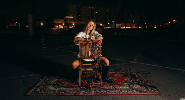 A woman sitting on a chair wrapped in roses, set on a carpet outdoors at night.