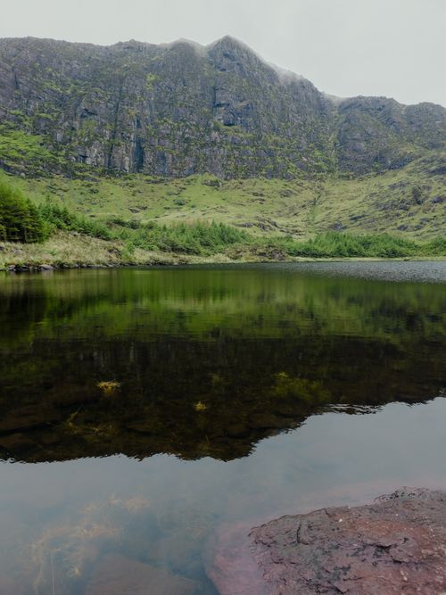 Free Tranquil Irish lake reflecting lush mountains and green landscape on a misty day. Stock Photo