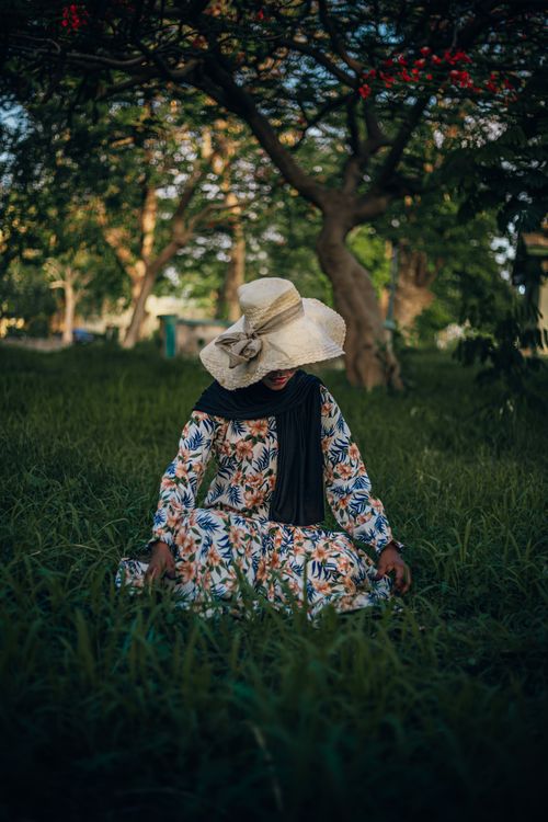 Free A woman in floral dress and wide-brimmed hat sits in a lush Nigerian garden. Stock Photo