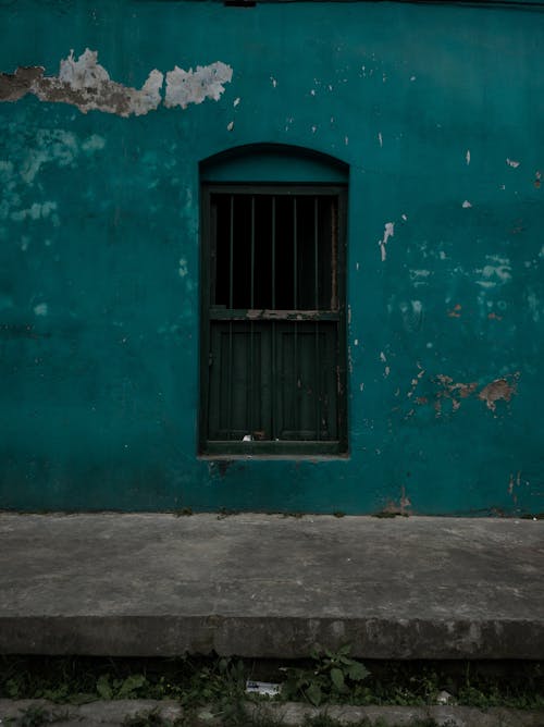 Free Exterior of abandoned building with rough walls with blots on surface and fenced rectangular window near cement pavement in evening Stock Photo