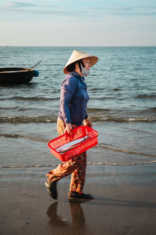 Free A woman walks along the beach holding a basket of fish, wearing a traditional conical hat. Stock Photo