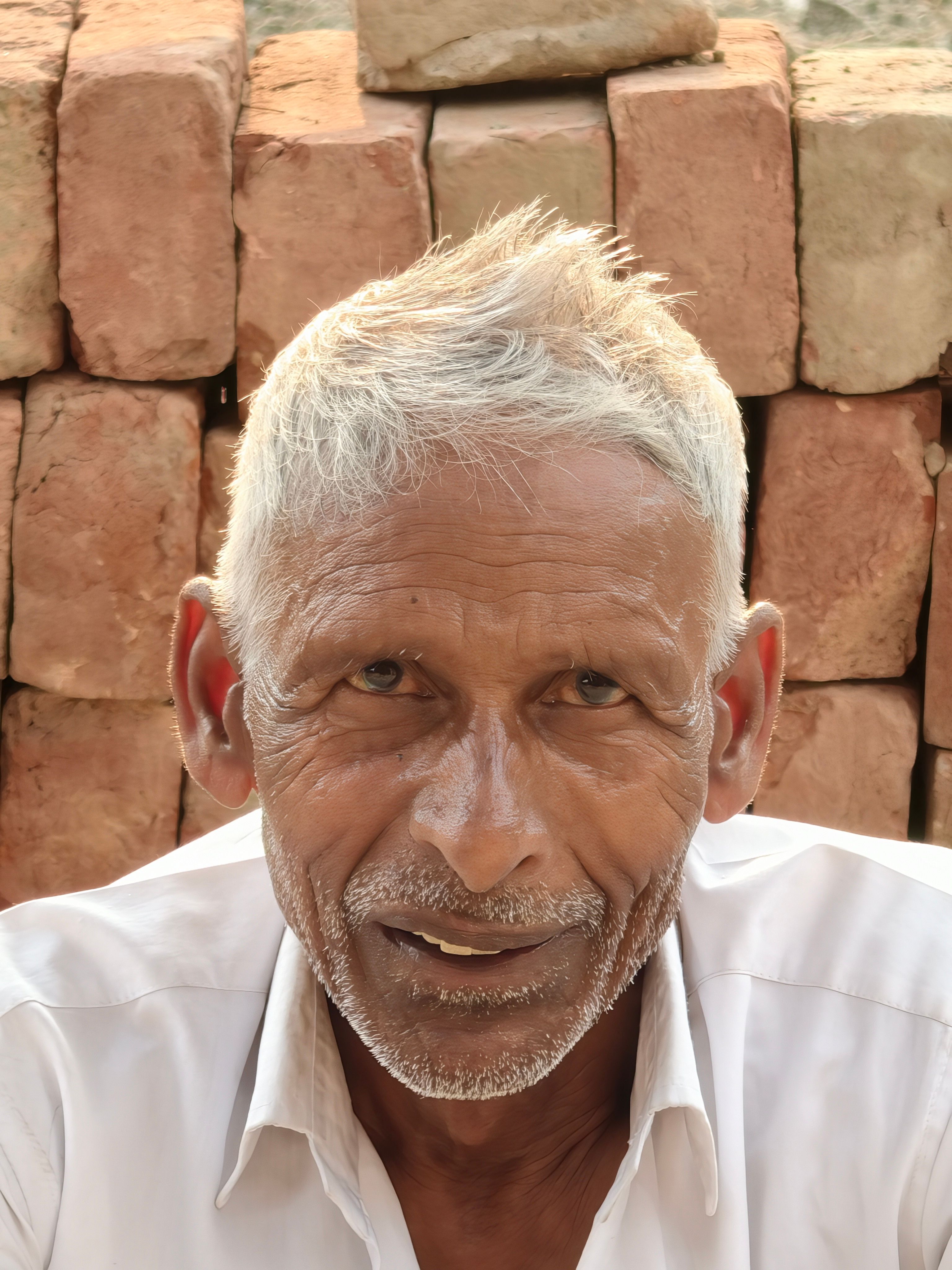 Portrait of an elderly man with white hair and a warm smile, sitting outdoors against a brick wall.