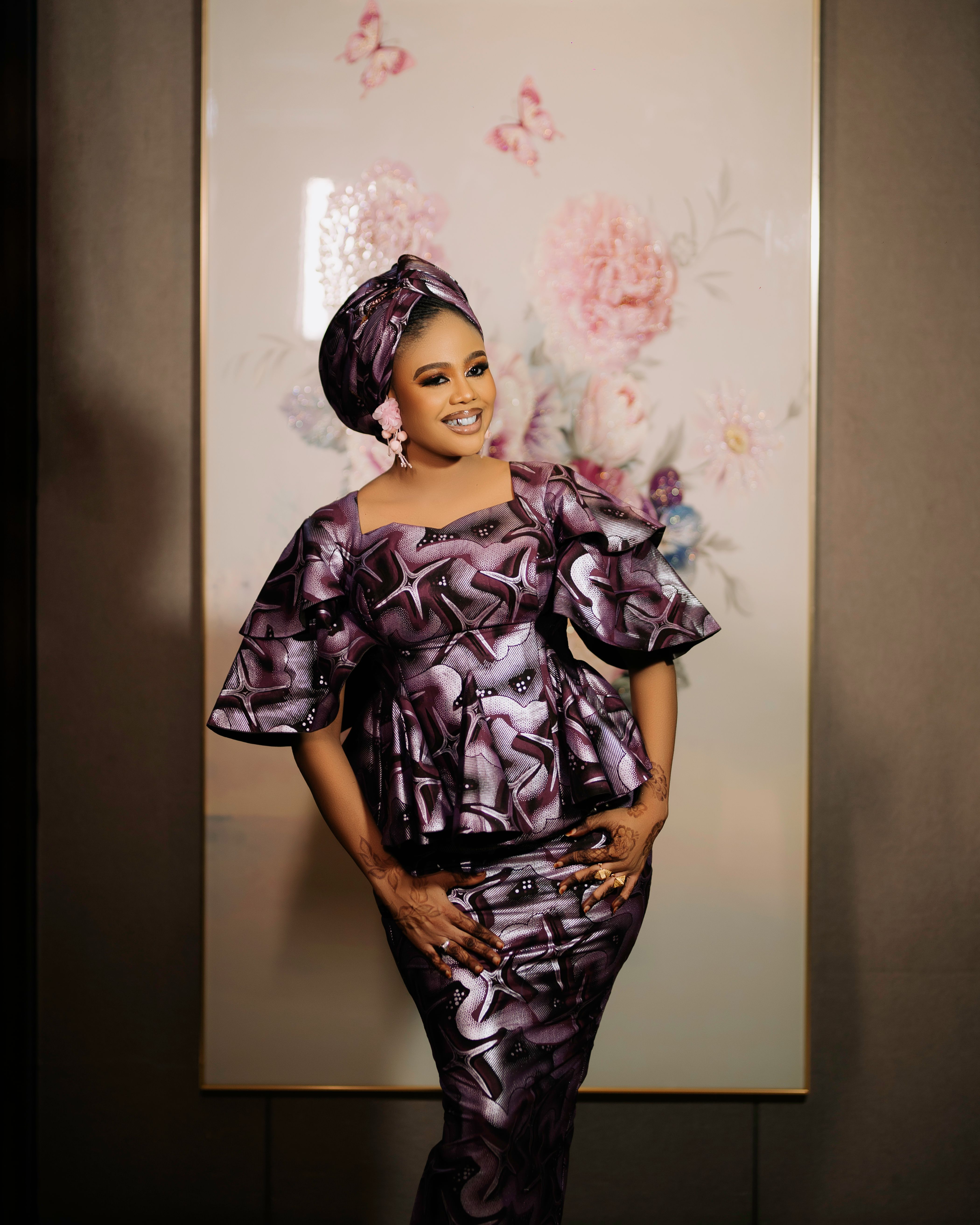 Woman in traditional African dress standing indoors against a floral art background.