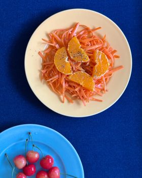 Vibrant salad of shredded carrots and oranges with cherries, against a blue background.