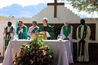Elderly priest in eyeglasses and catholic vestment standing with prayer hands near table with Bible and multiethnic deacons during liturgy process behind mountains in sunlight