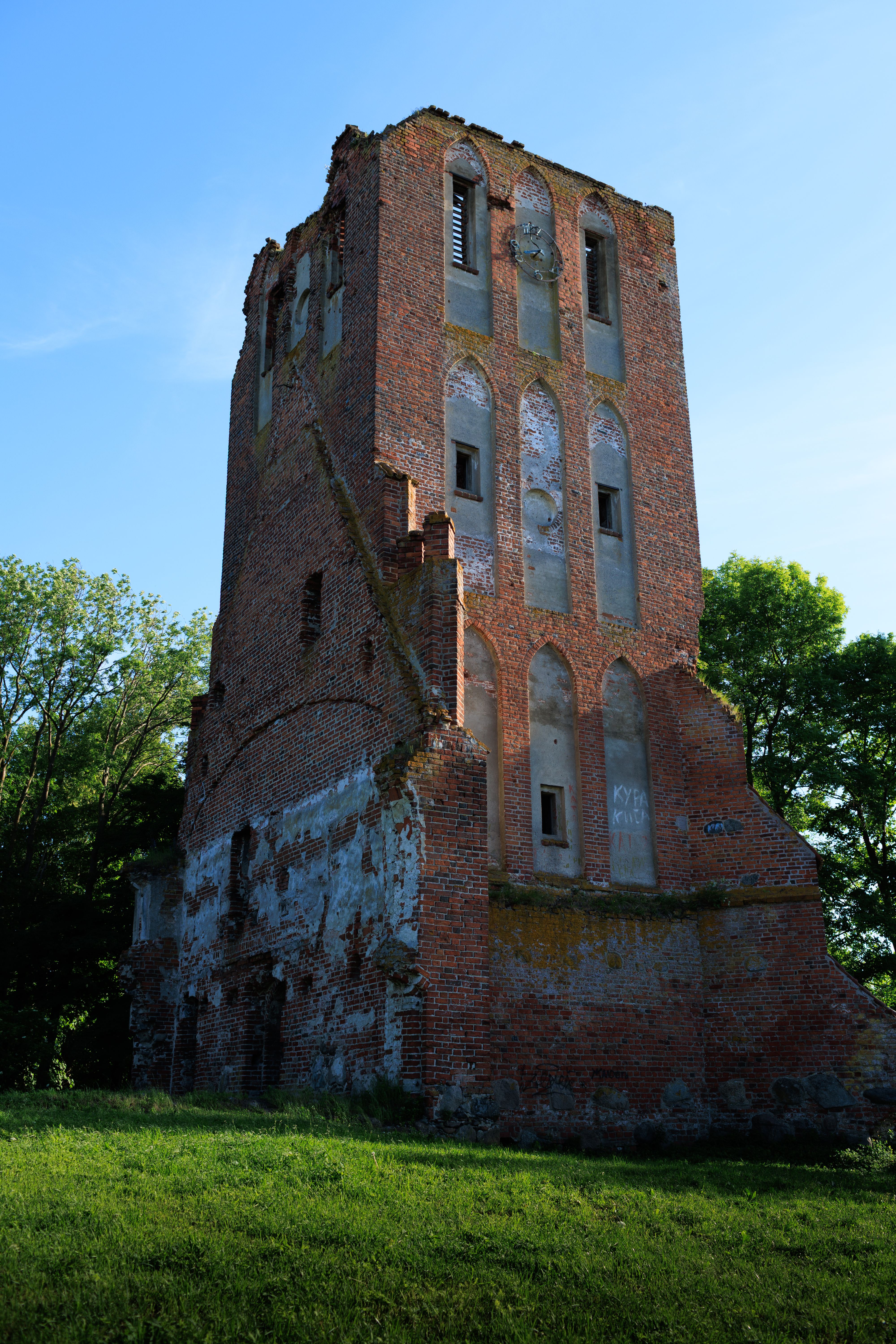 A rustic, abandoned brick tower stands amidst lush greenery under a clear blue sky.