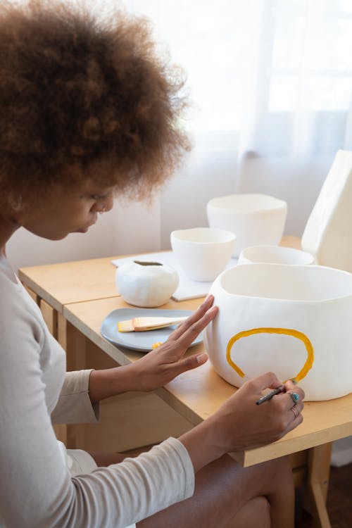 Free From above side view of talented African American female artisan sitting at table in creative workshop and paining picture on handmade ceramic bowl Stock Photo