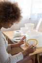 From above side view of talented African American female artisan sitting at table in creative workshop and paining picture on handmade ceramic bowl