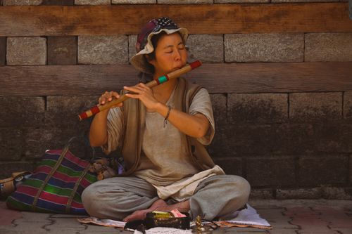 Free A street performer sits cross-legged playing a colorful wooden flute against a rustic brick wall. Stock Photo
