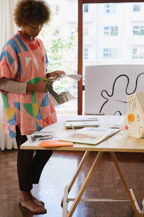 Creative black female craftswoman wearing trendy clothes standing near wooden table and making paper cutouts with scissors