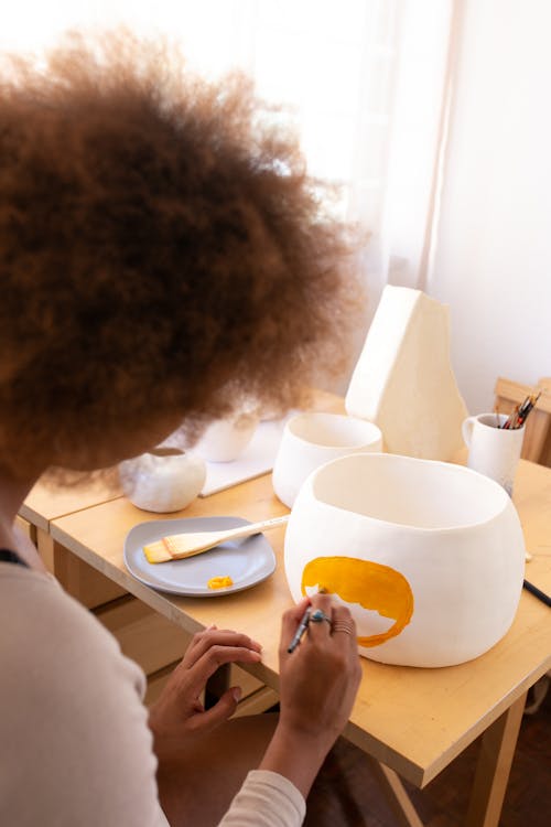 From above back view of crop ethnic female artisan with curly hair sitting at table in workshop and creating pictures with paintbrush on clay tableware