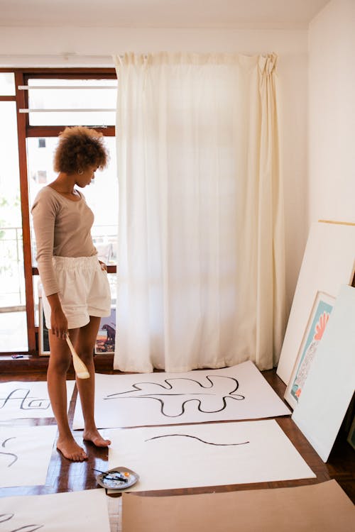 Side view of black barefoot female in casual outfit standing at cozy workspace and looking at colorful illustration by wall and abstract paintings on floor