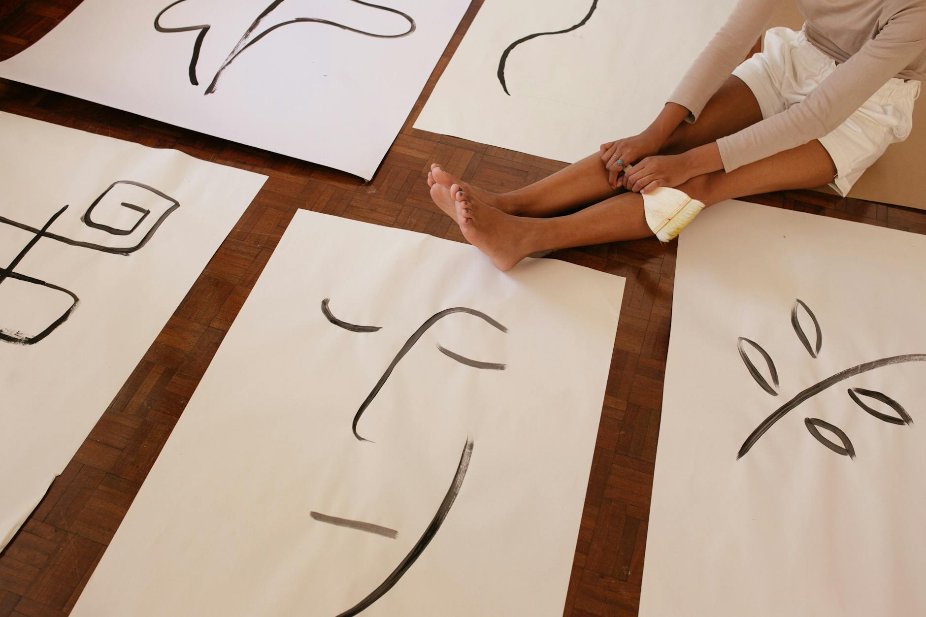 From above of faceless barefoot casually dressed woman with brush sitting on floor while placed artworks in abstract style