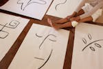 From above of faceless barefoot casually dressed woman with brush sitting on floor while placed artworks in abstract style