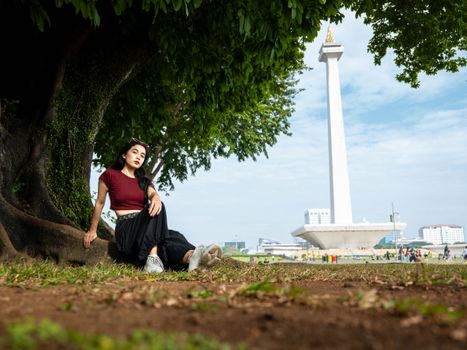 A young woman sits under a tree by Monas, Jakarta, enjoying a sunny day outdoors.