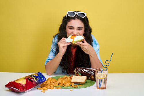 Free A cheerful young woman eating a sandwich with chips and a drink on a vibrant background. Stock Photo
