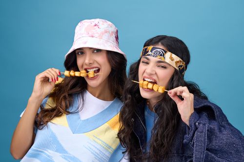 Free Two friends in stylish outfits happily enjoy skewered snacks against a vibrant backdrop. Stock Photo