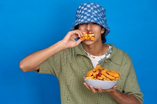 Free A stylish young adult eating fast food from a bowl, set against a vibrant blue backdrop. Stock Photo
