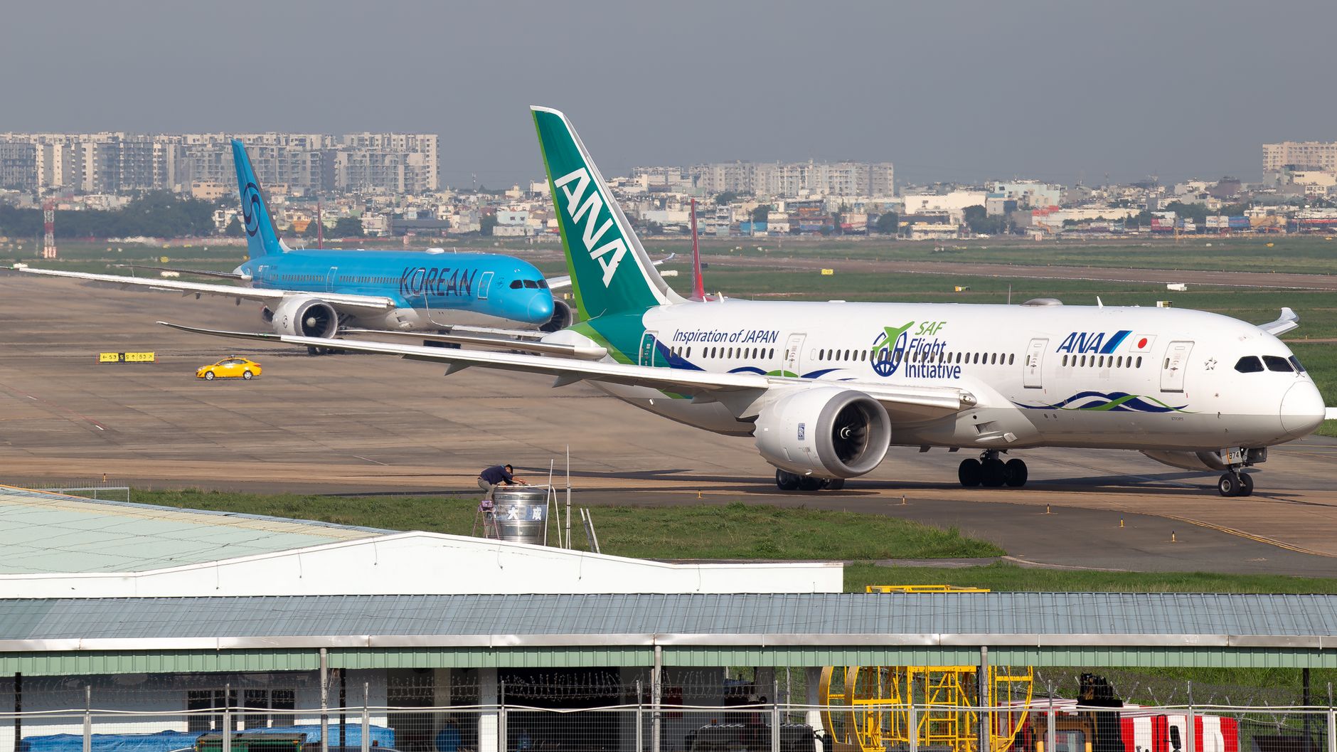 Two commercial airplanes taxi on a busy airport runway with cityscape in the background.