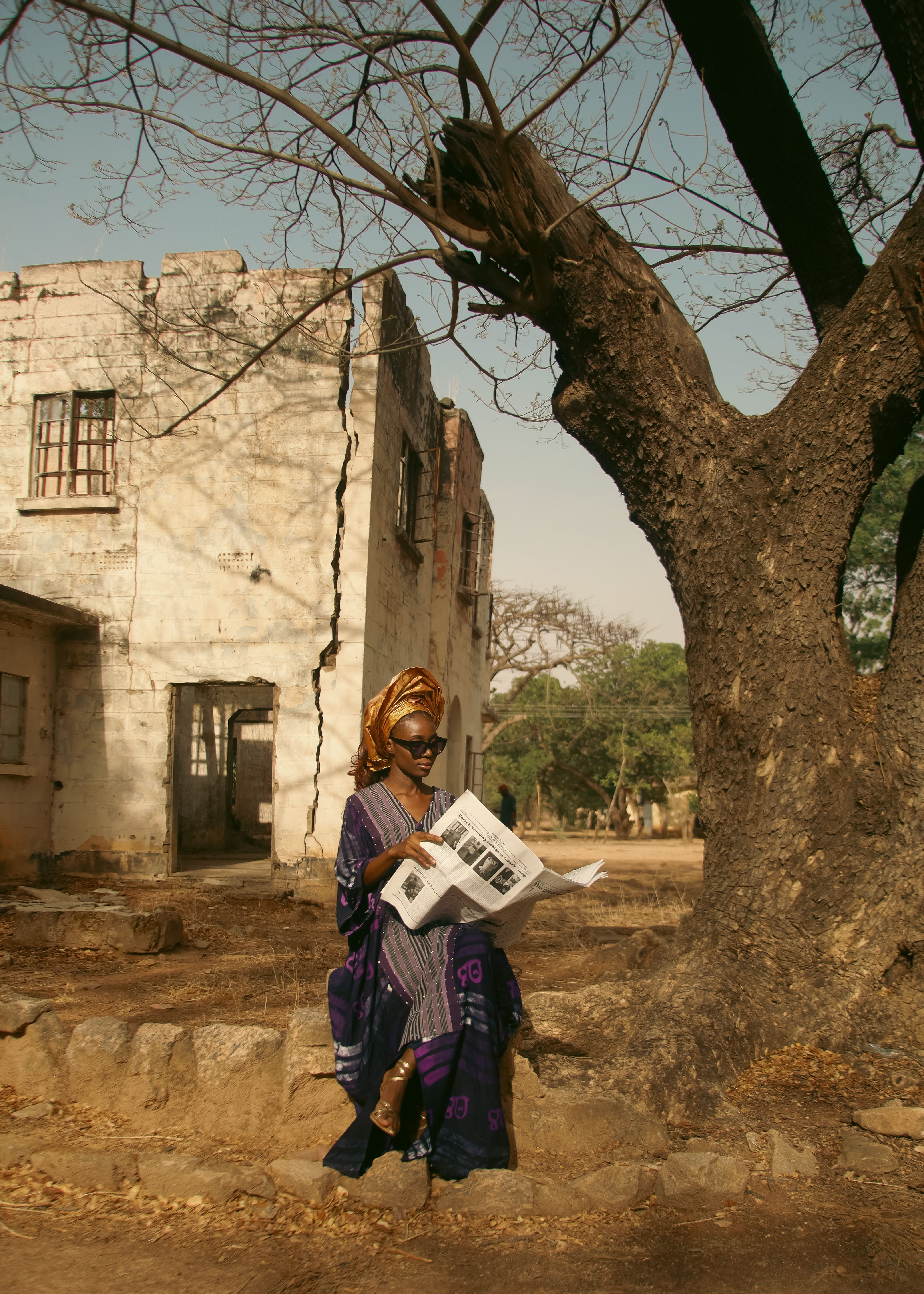 African woman in traditional attire reading a newspaper outdoors near an old building