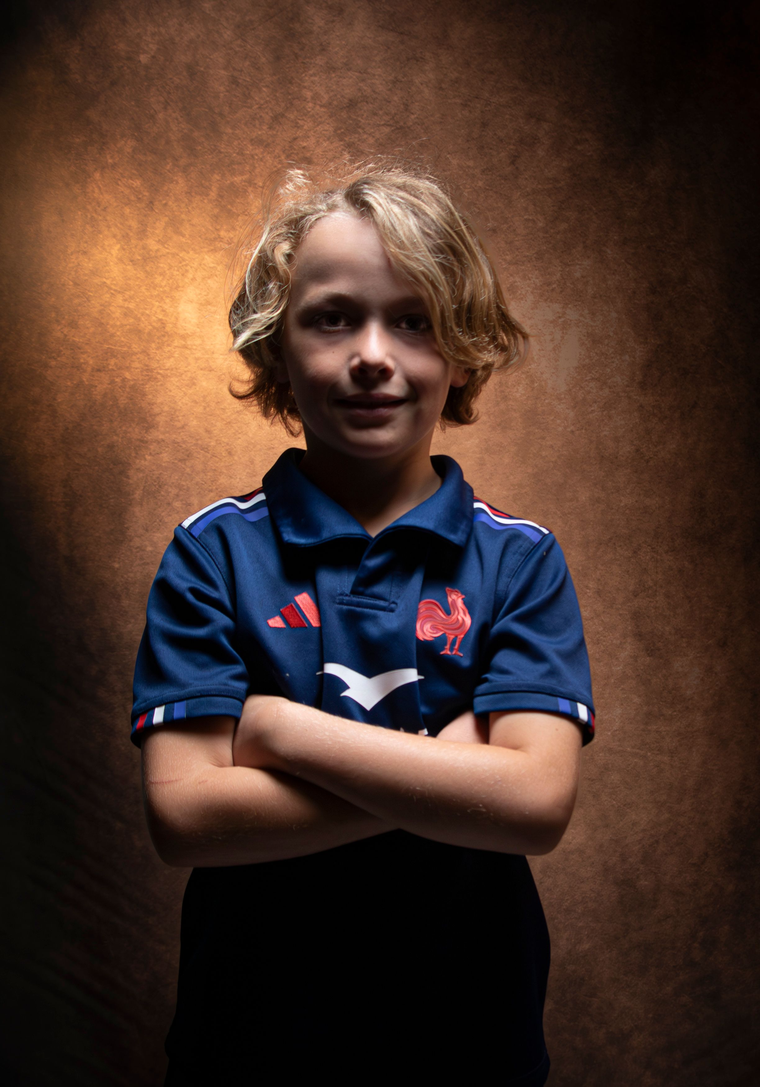 Smiling young boy wearing a blue sports jersey, arms crossed, dramatic lighting.