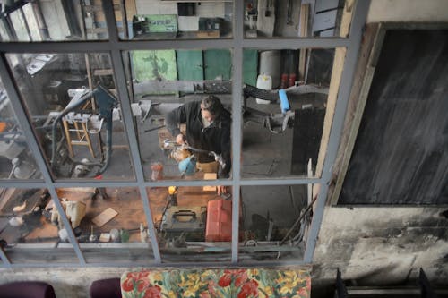 Through window view of male worker in casual uniform standing near workbench and working with instruments in grungy workshop