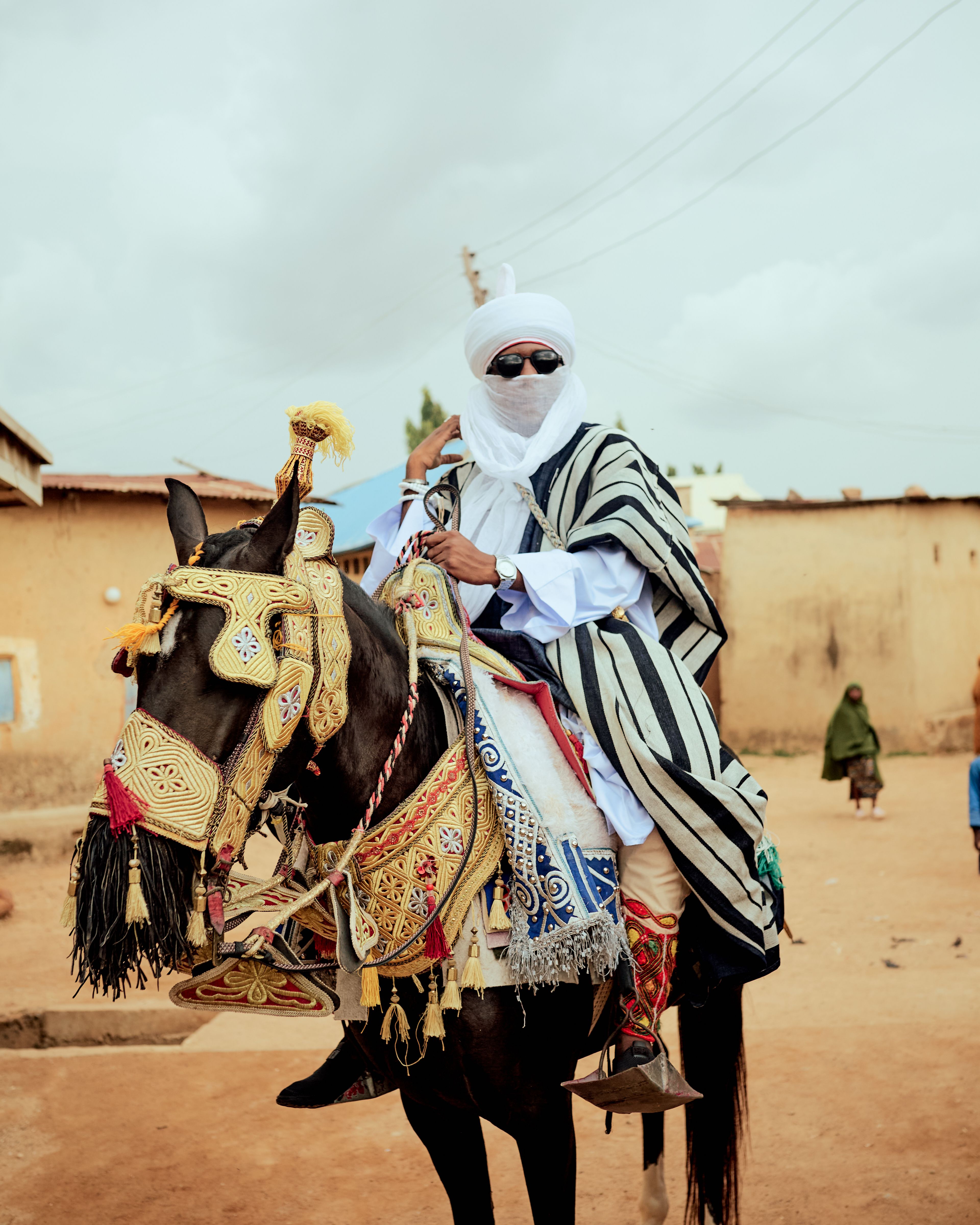 A Tuareg man dressed in traditional attire riding a richly adorned horse in a village setting.