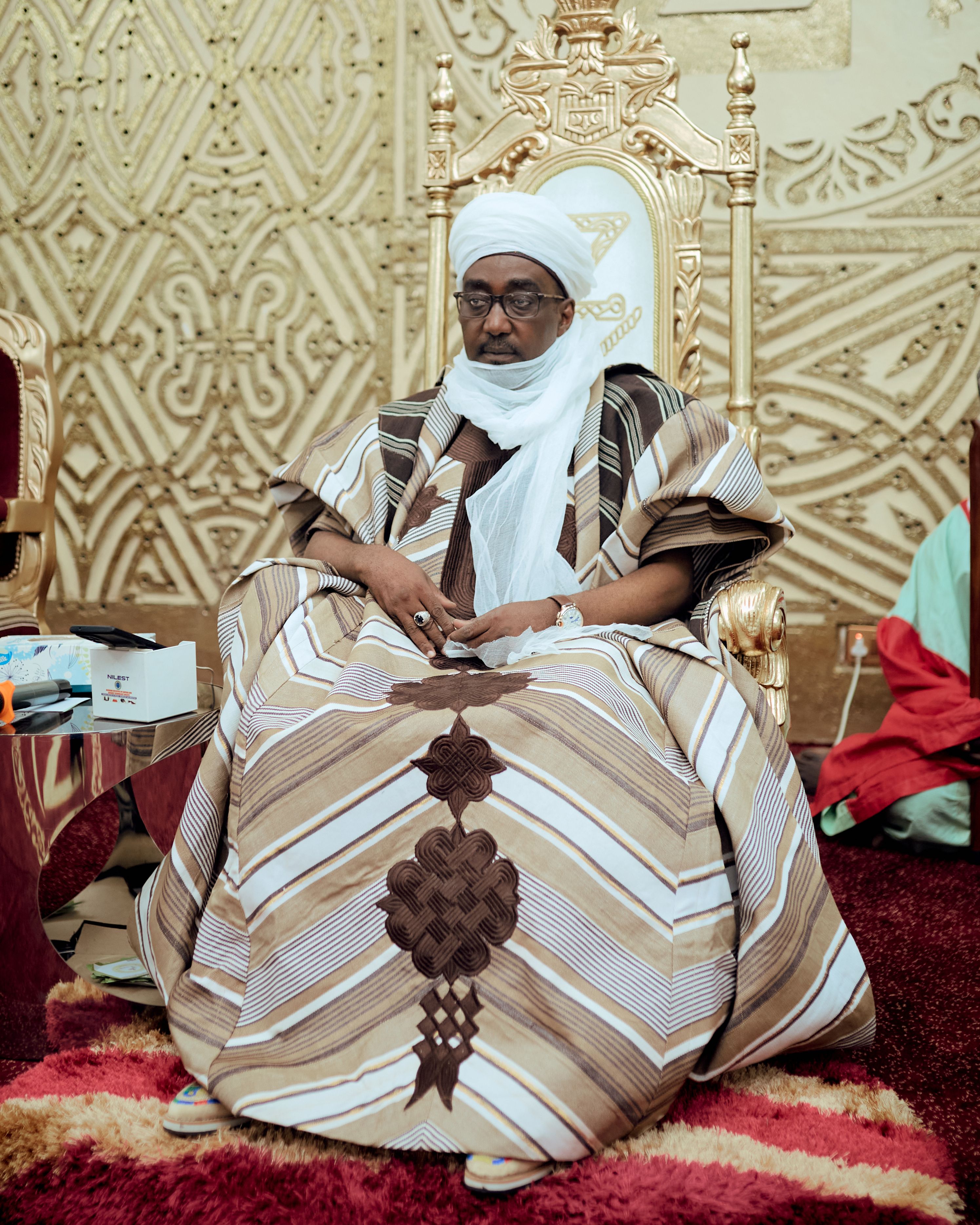 A traditional African leader in ceremonial attire seated on an ornate throne indoors.