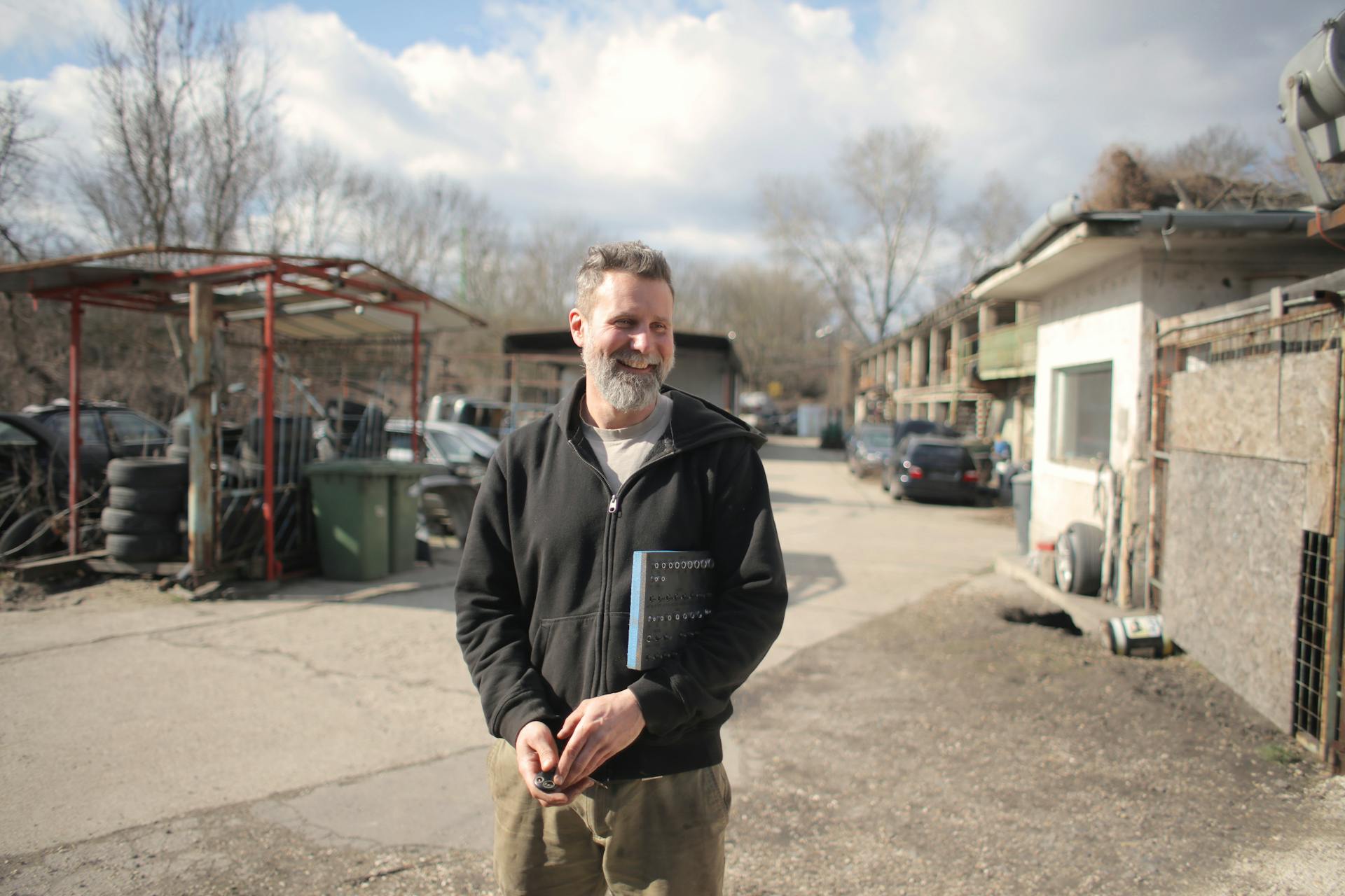 A man in a black jacket smiles while standing in an auto salvage yard on a sunny day.